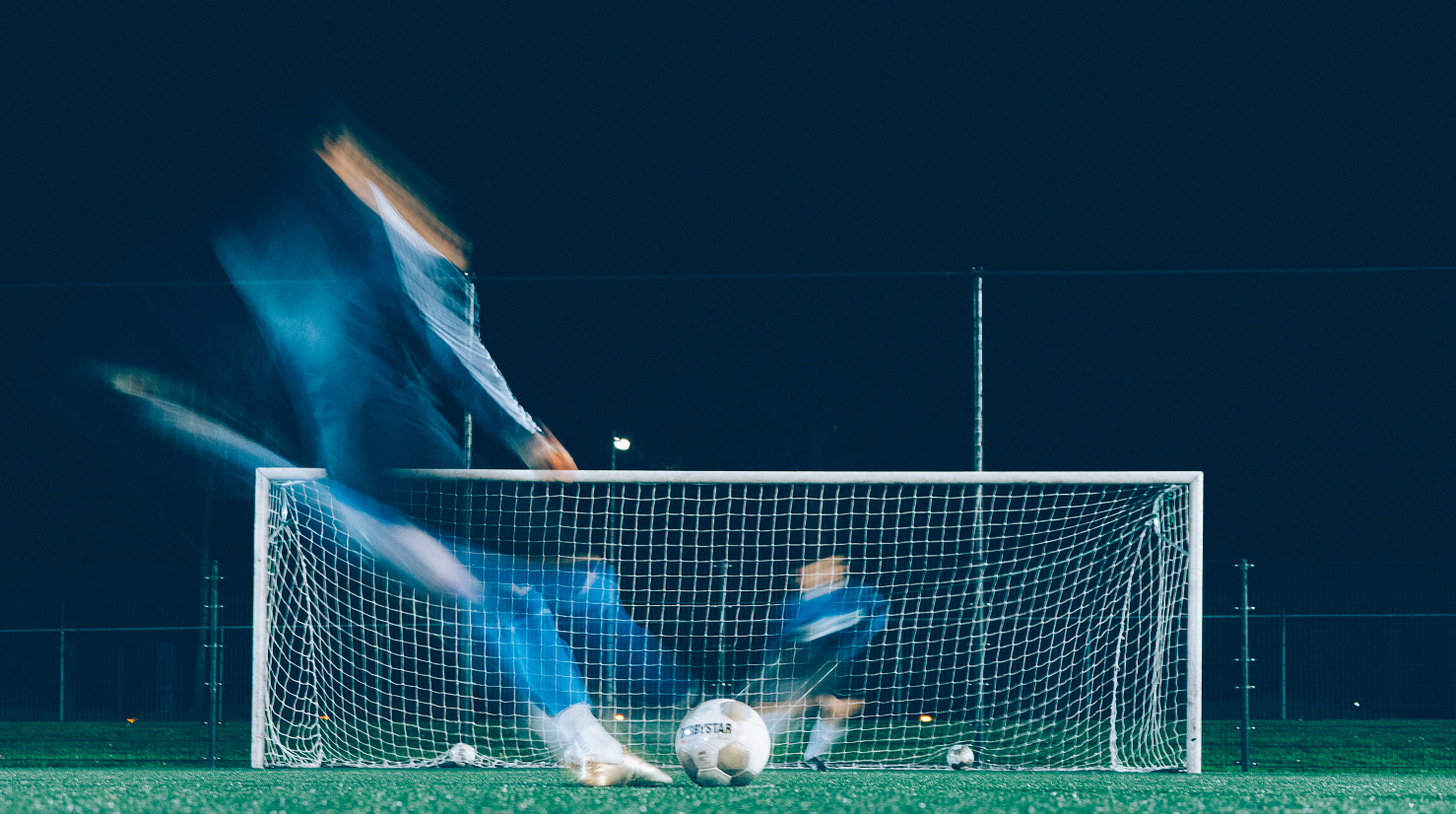 Color photo of two blurred figures on a football pitch at night, one kicking a ball and the other defending the goal
