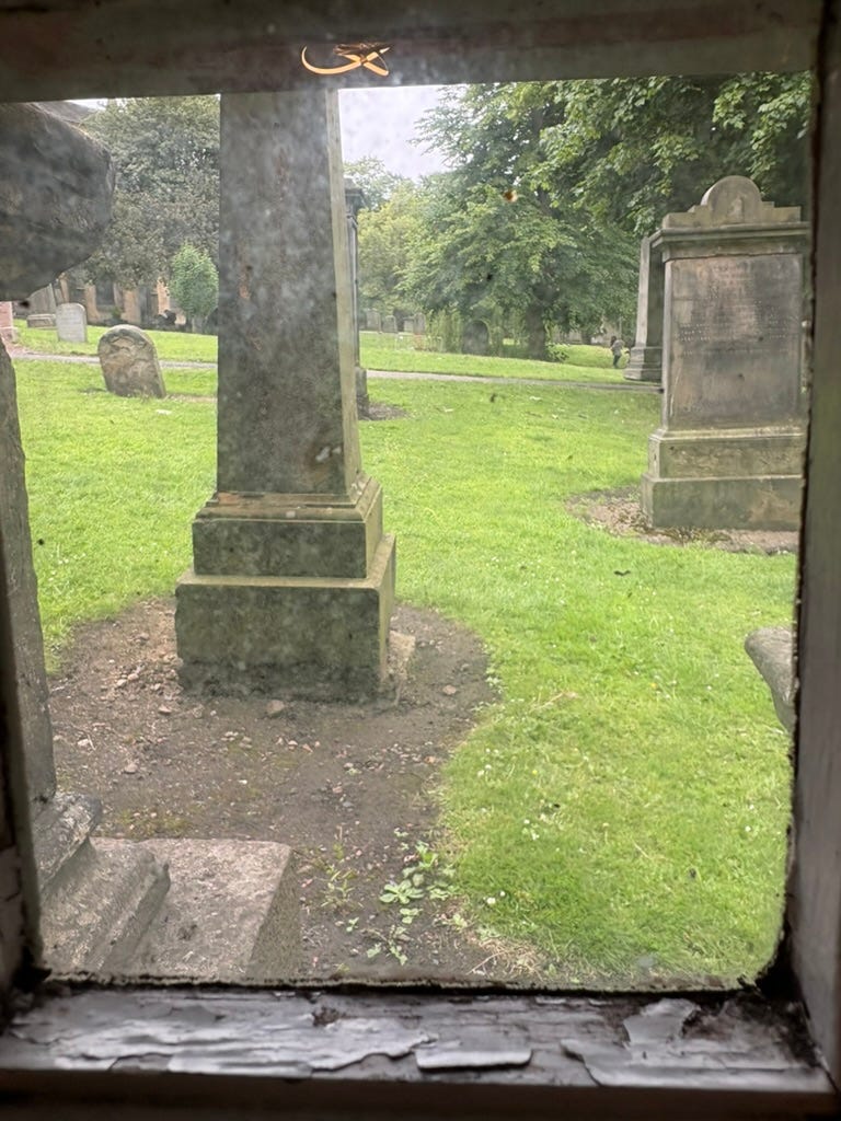 Photo looking from a window into a backyard that's actually a very old cemetery! To the immediate left of the window you can see part of a headstone literally up against the house! Other grave markers are visible against the green grass, stretching for a ways