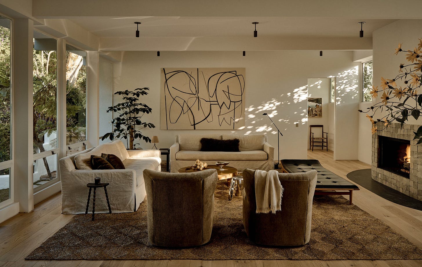 A warm, modern living room features large windows, neutral furniture, abstract art, a cozy fireplace, indoor plants, and soft natural light illuminated by the Long Made Co. Brass Cylinder Drop Spot - Top Mount ceiling downlight casting gentle shadows.
