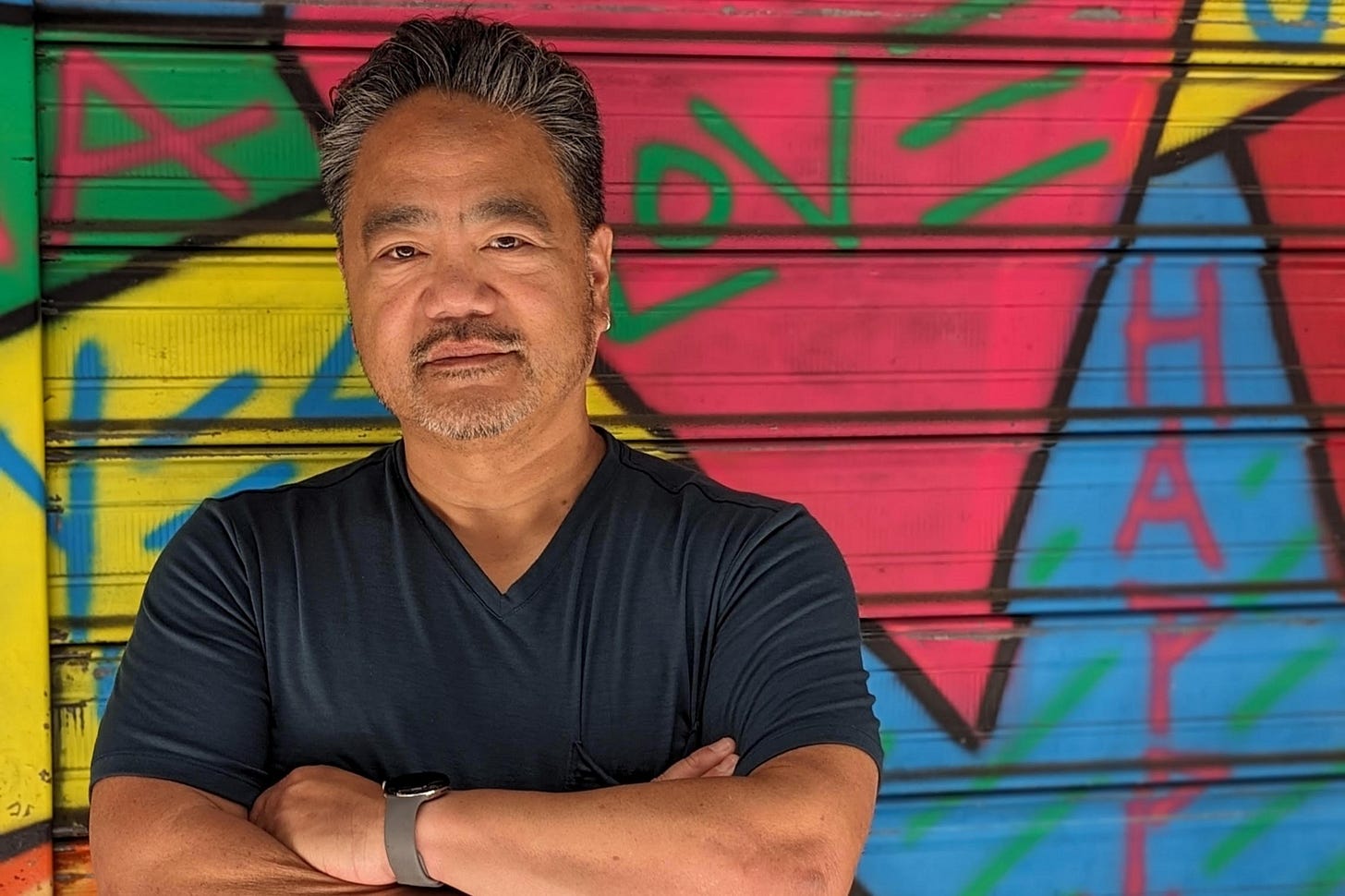 Bruce, a middle aged Asian American man with salt-and-pepper hair and a light beard and mustache looks directly at the camera. He wears a black T shirt and his arms are folded across his chest. Behind him is a colorful graffitti wall with the word LOVE.