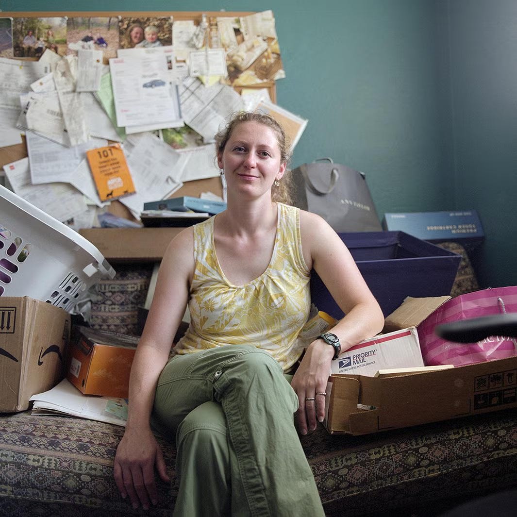 woman on cluttered couch, box beside her, cluttered bulletin board behind her; she's wearing short sleeve yellow blouse and greenish trousers woman on cluttered couch, box beside her, cluttered bulletin board behind her; she's wearing short sleeve yellow blouse and greenish trousers
