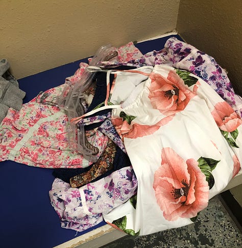 A pile of floral dresses teeters on the thrift store bench. Four are chosen: a goldenrod, a white-with-purple, a midnight blue with a lace cutout at the waistband, and a multi-hued jaggedy pattern with puffy cap sleeves.
