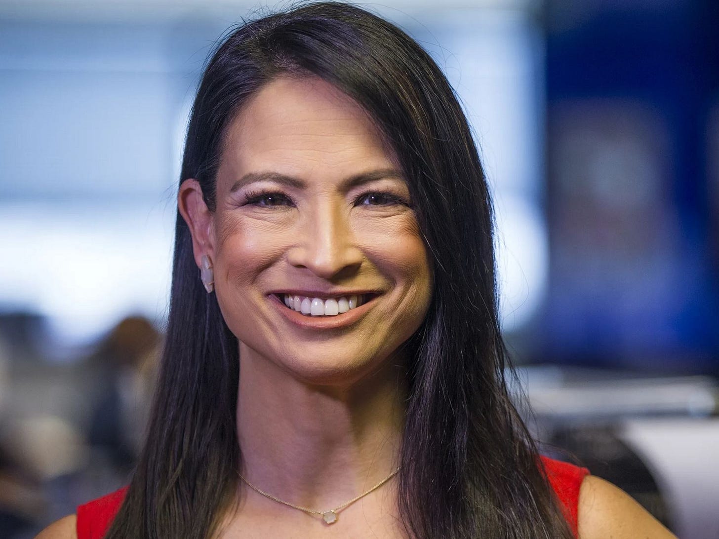 An Asian-American woman with long, dark hair smiles confidently in a professional setting. She is wearing a red outfit and a delicate necklace, with a blurred newsroom background behind her.