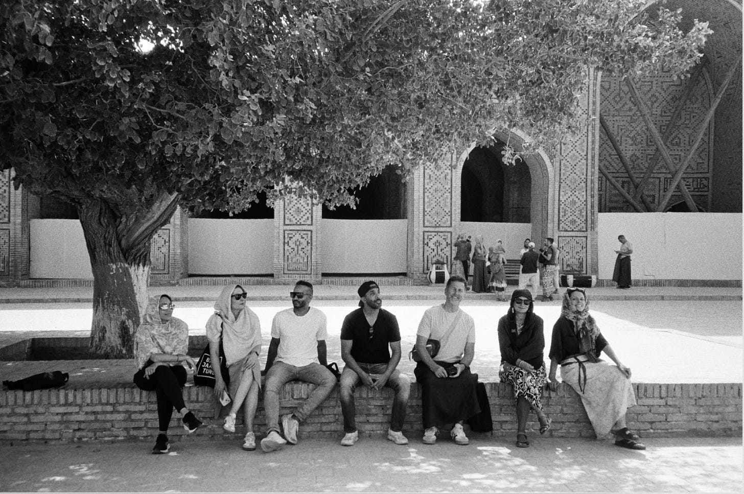 Black and white photo of 7 tourists sitting on a brick wall outside a Central Asian tiled temple. Black and white photo of 7 tourists sitting on a brick wall outside a Central Asian tiled temple.