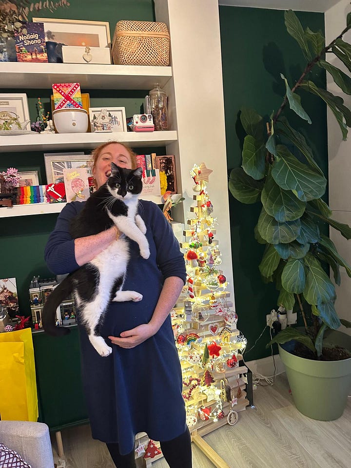 A pregnant woman (& her cat) in front of a wooden Christmas tree and some shelves