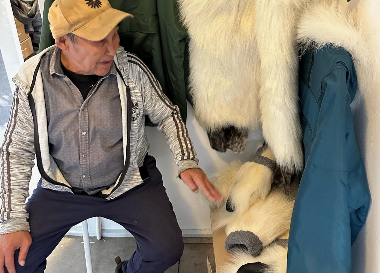 Local man sitting beside white polar bear fur garments hanging on the wall in a small Greenland shop.