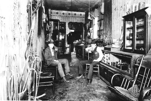 Interior of H. Heines Harness Shop: Rows of bridles, collars, and straps hang neatly along the walls as Henry and a co-worker sit.(Photo from Lynda Heines family collection)