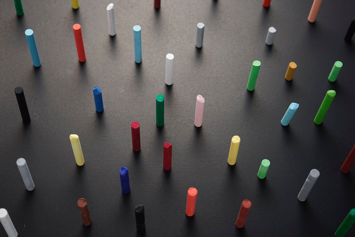 Superficie grigio-nera su cui sono appoggiati in verticale e l'uno distante dall'altro tanti pastelli a cera di colori diversi. Superficie grigio-nera su cui sono appoggiati in verticale e l'uno distante dall'altro tanti pastelli a cera di colori diversi.