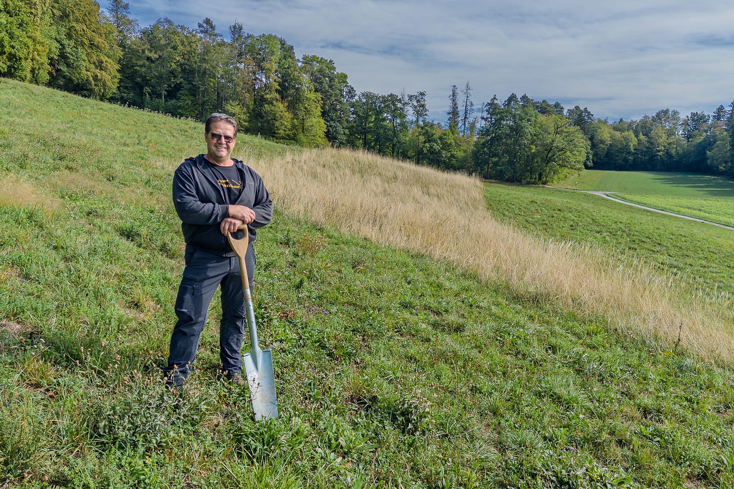 Landwirt Armon Fliri steht mit einem Pflanzspaten in einer extensiv bewirtschafteten Wiese am Sonnenberg. Hinter ihm zieht sich ein breiter Altgrasstreifen mit hohem, trockenem Gras durch die grüne Hangwiese; im Hintergrund liegen Wald und weitere Landwirtschaftsflächen. Landwirt Armon Fliri steht mit einem Pflanzspaten in einer extensiv bewirtschafteten Wiese am Sonnenberg. Hinter ihm zieht sich ein breiter Altgrasstreifen mit hohem, trockenem Gras durch die grüne Hangwiese; im Hintergrund liegen Wald und weitere Landwirtschaftsflächen.