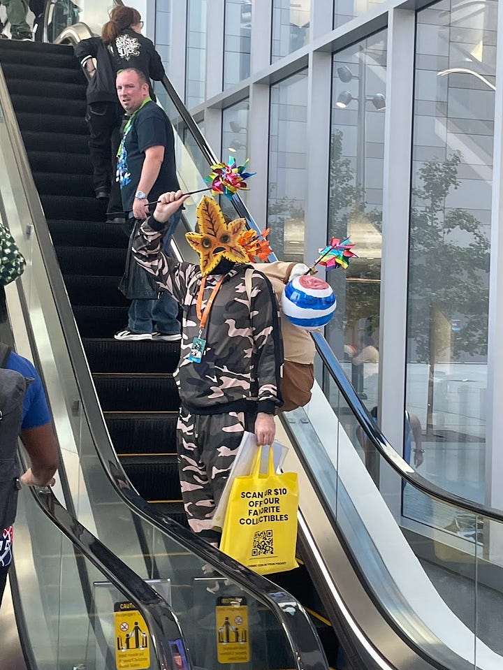Lauren poses with a thumbs up next to a man cosplaying as a Korok in camo pants and Korok mask. Another man in Korok cosplay waving from an escalator.