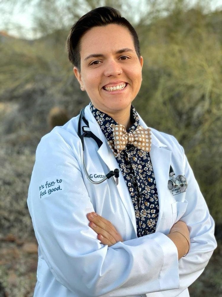person with short dark hair is wearing a white lab coat, dark blue floral button up, and gold bow tie. they have a stethescope around their neck and are smiling. this is dr. sg gettman, nd. 