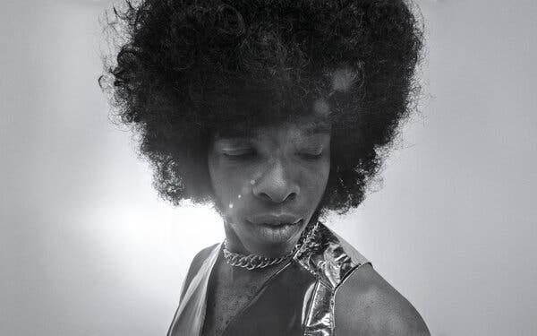 A man with an Afro and a gold chain and vest looks downward. 