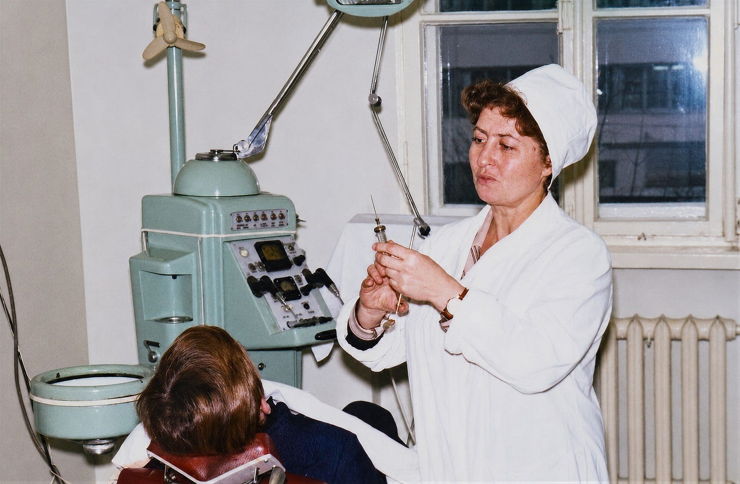 A nurse in a white uniform prepares a syringe next to a seated patient in a medical clinic with vintage equipment and large windows.