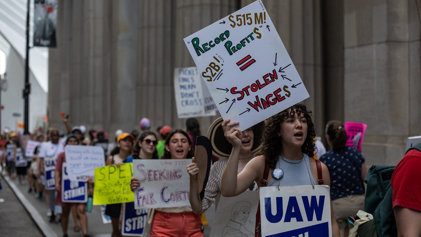 Image by Benjamin Norman for the New York Times; a photograph of Harper Collins workers striking outside the Harper offices, holding many picket signs. 