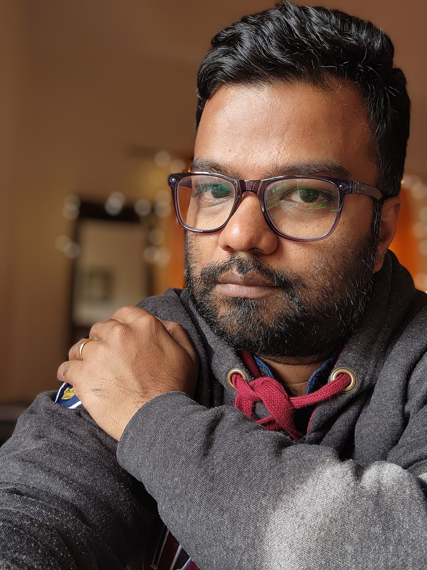 Tanmoy, an Indian man with dark hair, a beard, and glasses, sits indoors, looking directly at the camera with a calm expression. He is wearing a dark gray hoodie with red drawstrings and has his left hand resting on his right shoulder.