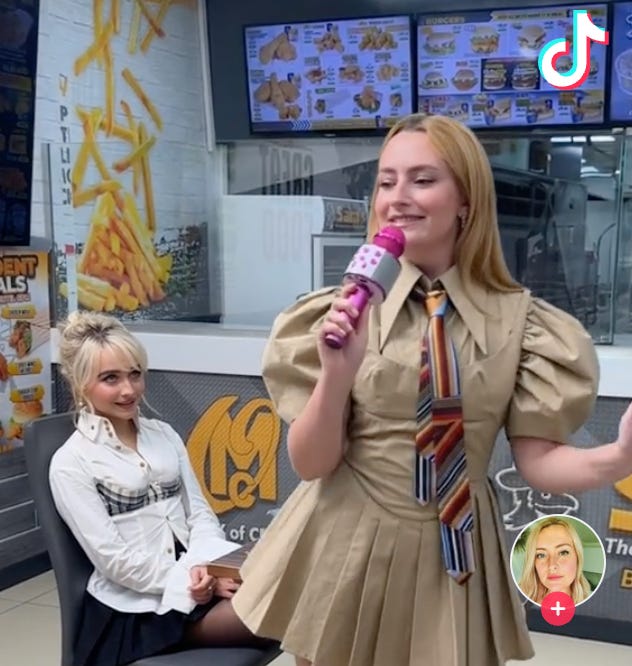 Sabrina Carpenter sits with a fake smile while watching Amelia from Chicken Shop Date lip sync to her song Espresso