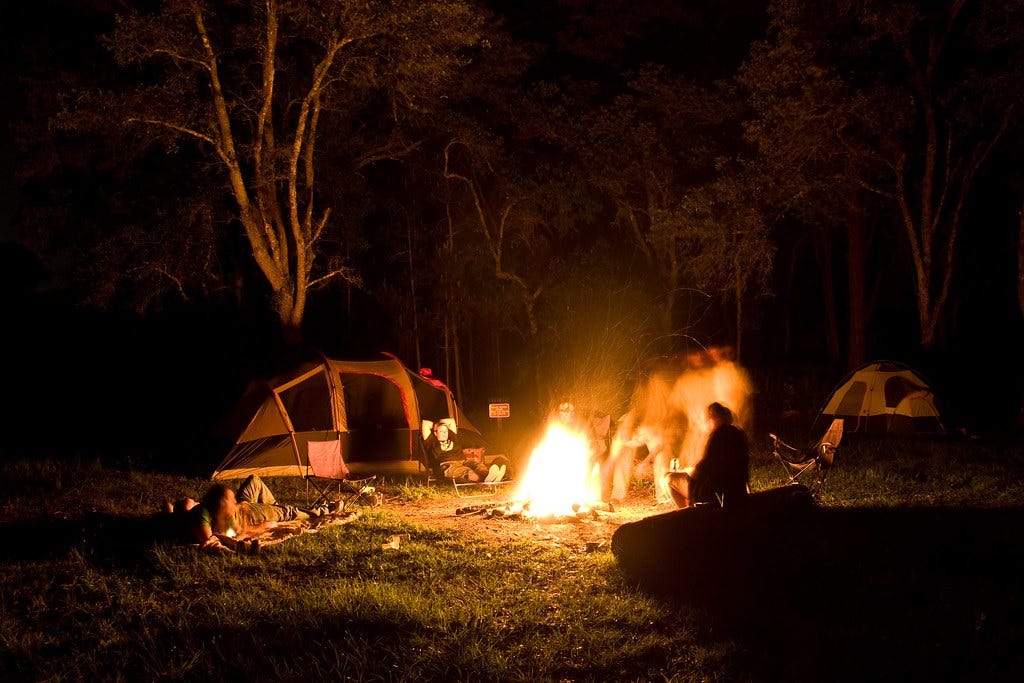 Photo of people sitting around a campfire that's burning brightly; one person stood up during the exposure, creating the appearance of a blurry 'ghost' Photo of people sitting around a campfire that's burning brightly; one person stood up during the exposure, creating the appearance of a blurry 'ghost'