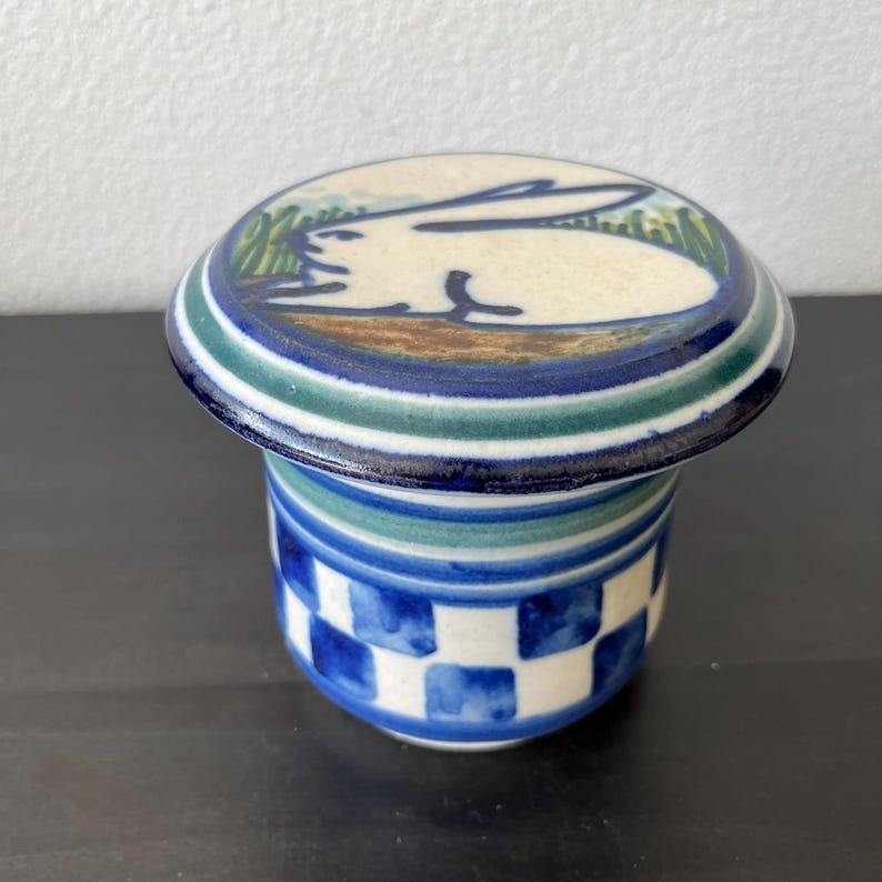 May include: A ceramic butter dish with a lid. The lid is round with a white rabbit design on top, surrounded by blue, green, and brown stripes. The base is cylindrical with a blue and white checkered pattern.