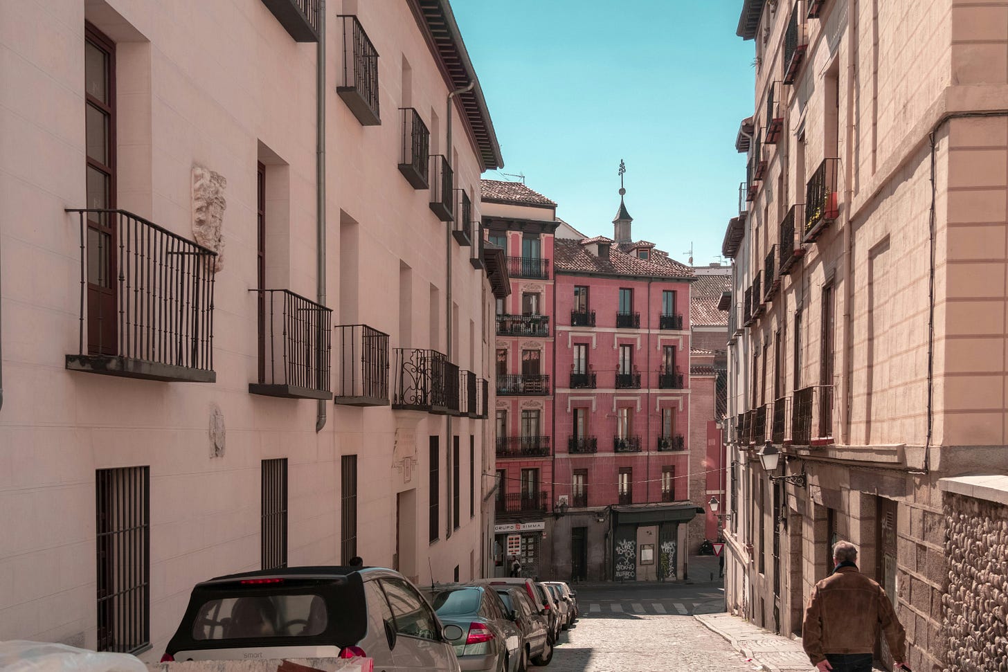 vehicles parked next to buildings in Madrid, Spain, with a man walking away from the camera down the cobblestone street
