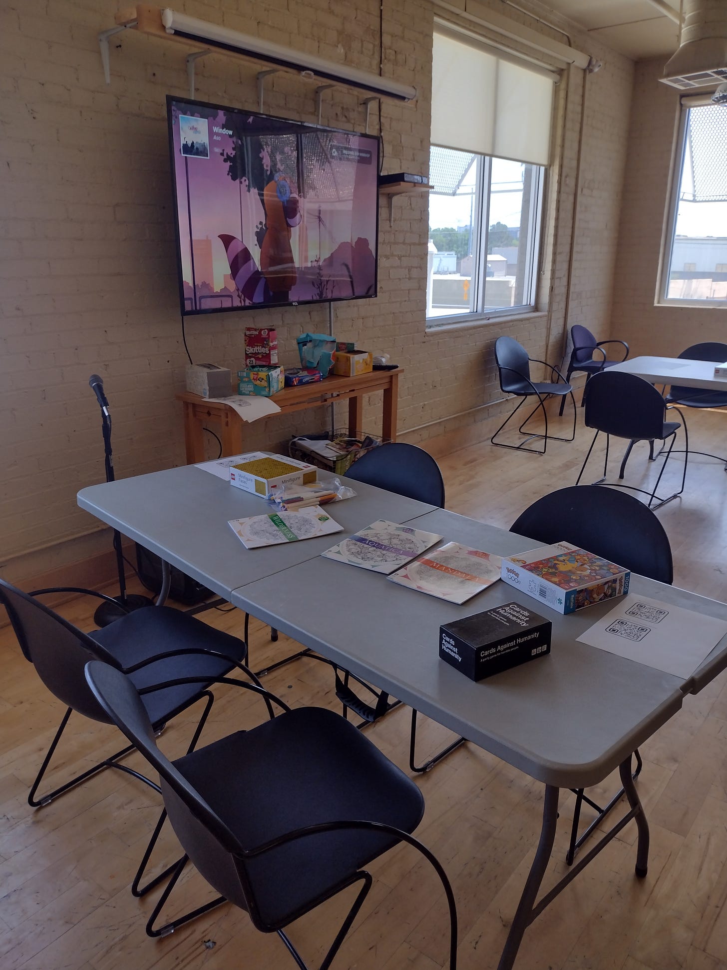 A table with four chairs and card games, puzzles, and coloring books on top of it. There is a wall in the background with a screen on it.
