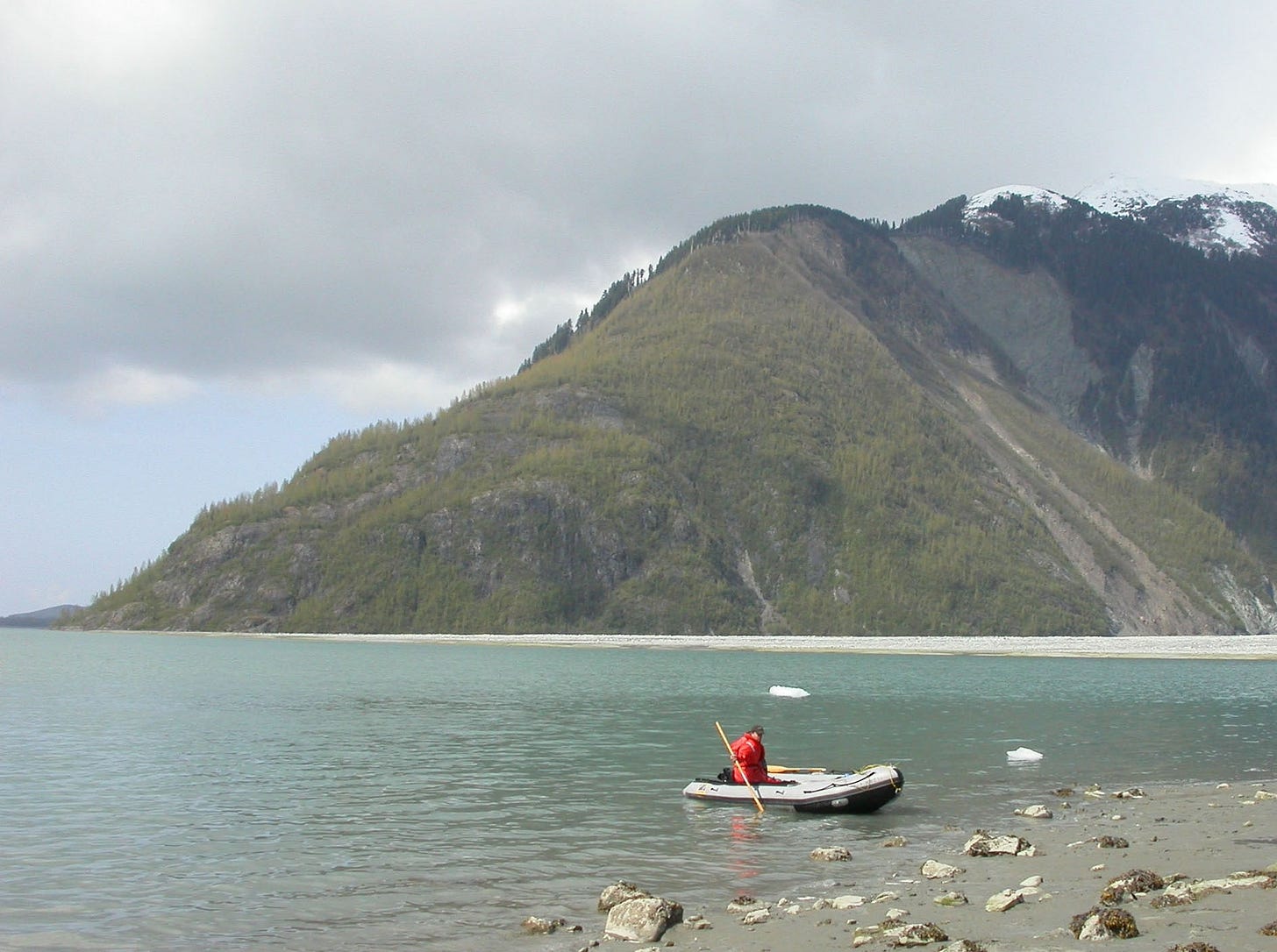 A person wearing a red flotation coat sits in an inflatable boat while bracing an oar in the water near a rocky shoreline. A few ice chunks float nearby. In the background, a steep, snow-topped mountain rises from the waterline. A person wearing a red flotation coat sits in an inflatable boat while bracing an oar in the water near a rocky shoreline. A few ice chunks float nearby. In the background, a steep, snow-topped mountain rises from the waterline.