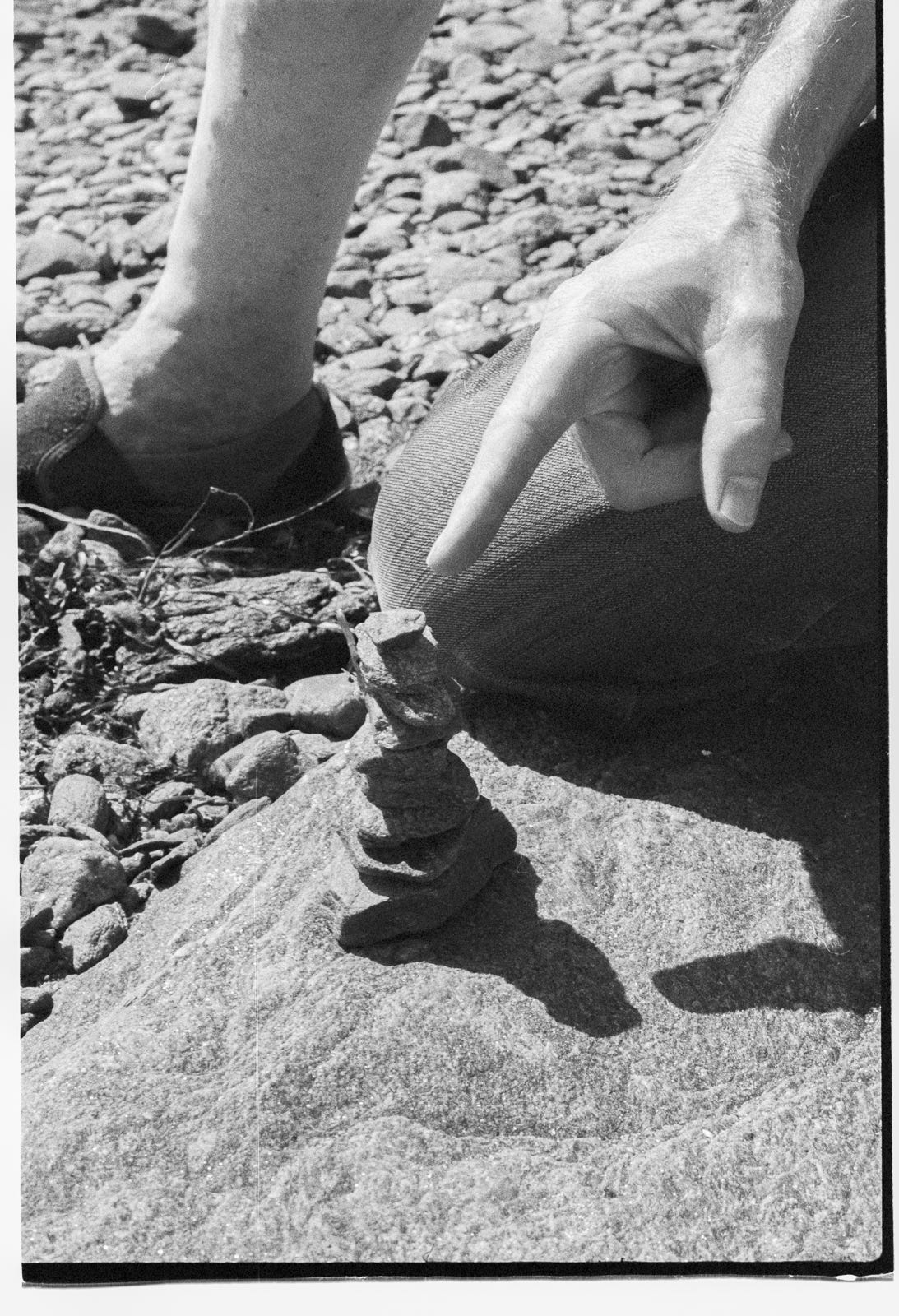 A hand points at a small stack of rocks on a beach. The owner of the hand is kneeling and you can see his knee and one of his feet. A hand points at a small stack of rocks on a beach. The owner of the hand is kneeling and you can see his knee and one of his feet.