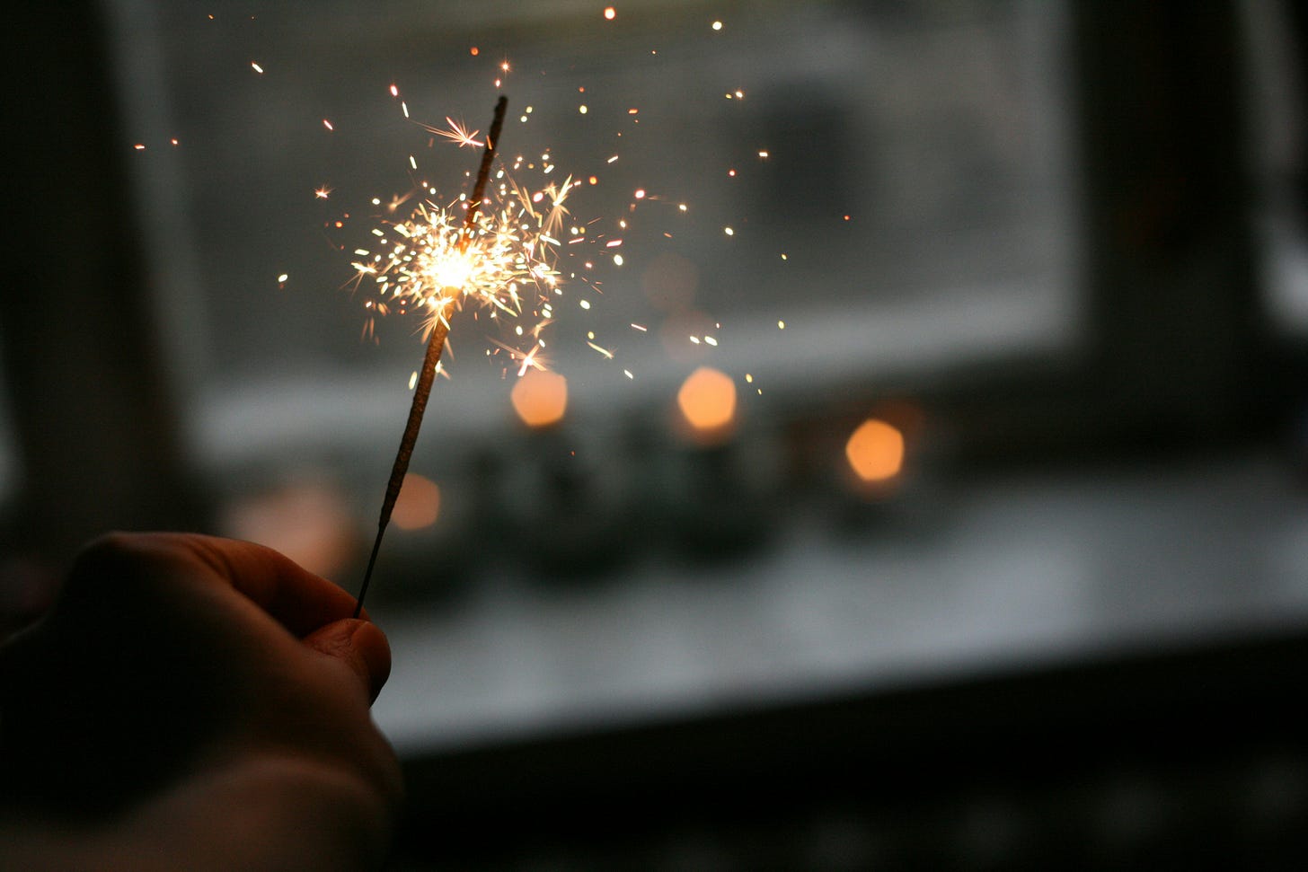 A hand holding a lit sparkler, a blurred laptop in the background A hand holding a lit sparkler, a blurred laptop in the background