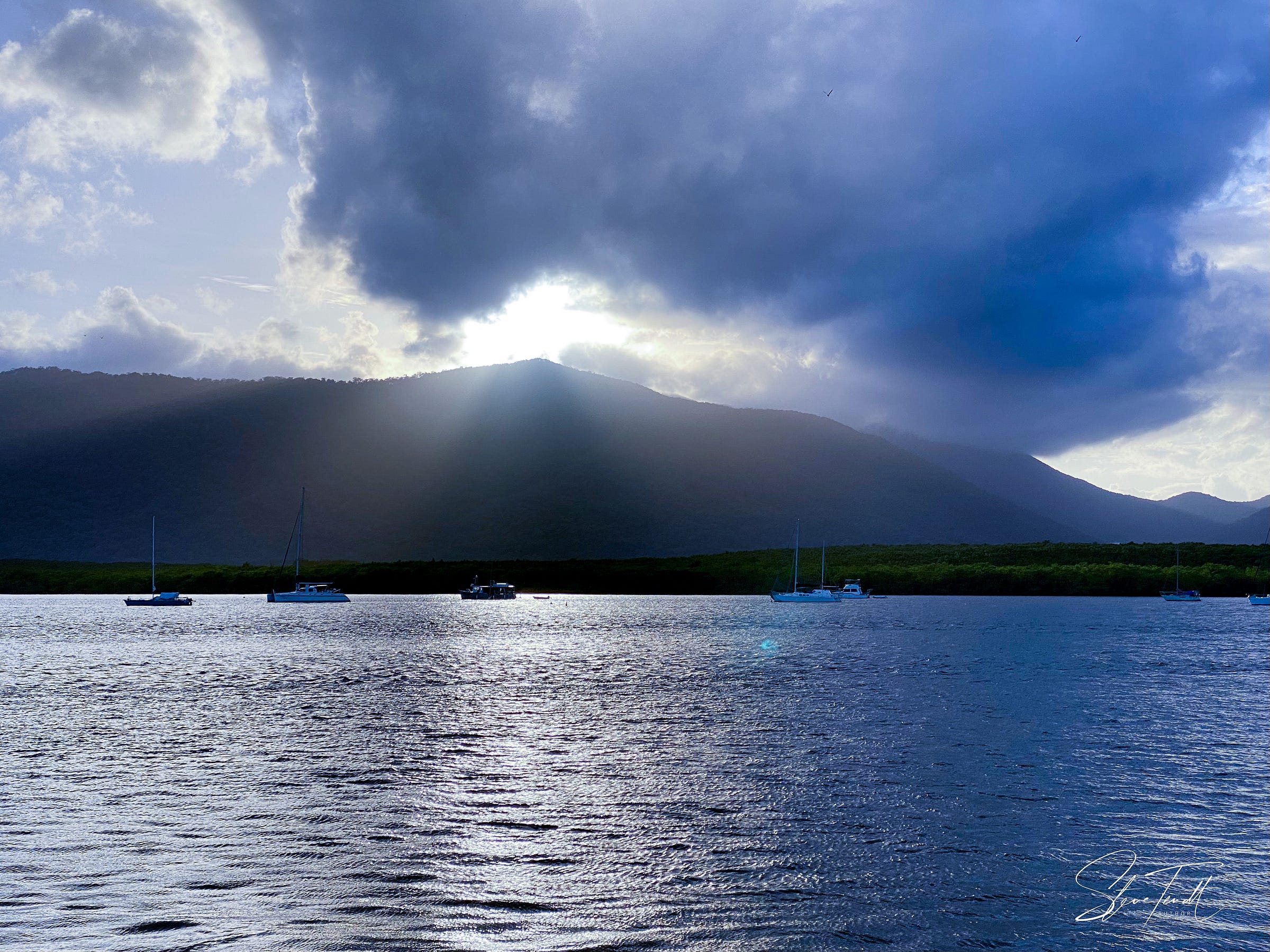 Photograph: strongly backlit landscape: Trinity Inlet, Cairns, with the water of the inlet in the foreground, yachts lying on their moorings in the distance, and behind the dark peak of a forest-covered mountain. Storm clouds are gathering over the peak, but the sun is trying to break through.