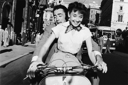 Audrey Hepburn rides a vespa with Gregory Peck behind her. A scene from Roman Holiday.