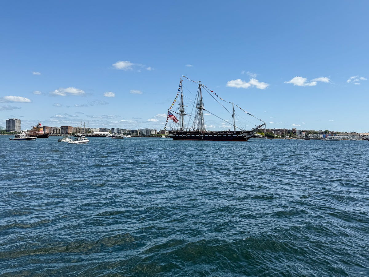 Wide shot of USS Constitution sailing with escort boats in Boston Harbor under clear blue sky Wide shot of USS Constitution sailing with escort boats in Boston Harbor under clear blue sky