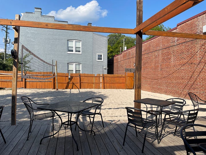 two photos of the same sand volleyball court with brick wall on right of photo and wooden picket fence along background; photo on left shows a platform deck with tables; photo on right shows to women looking up to watch for the ball