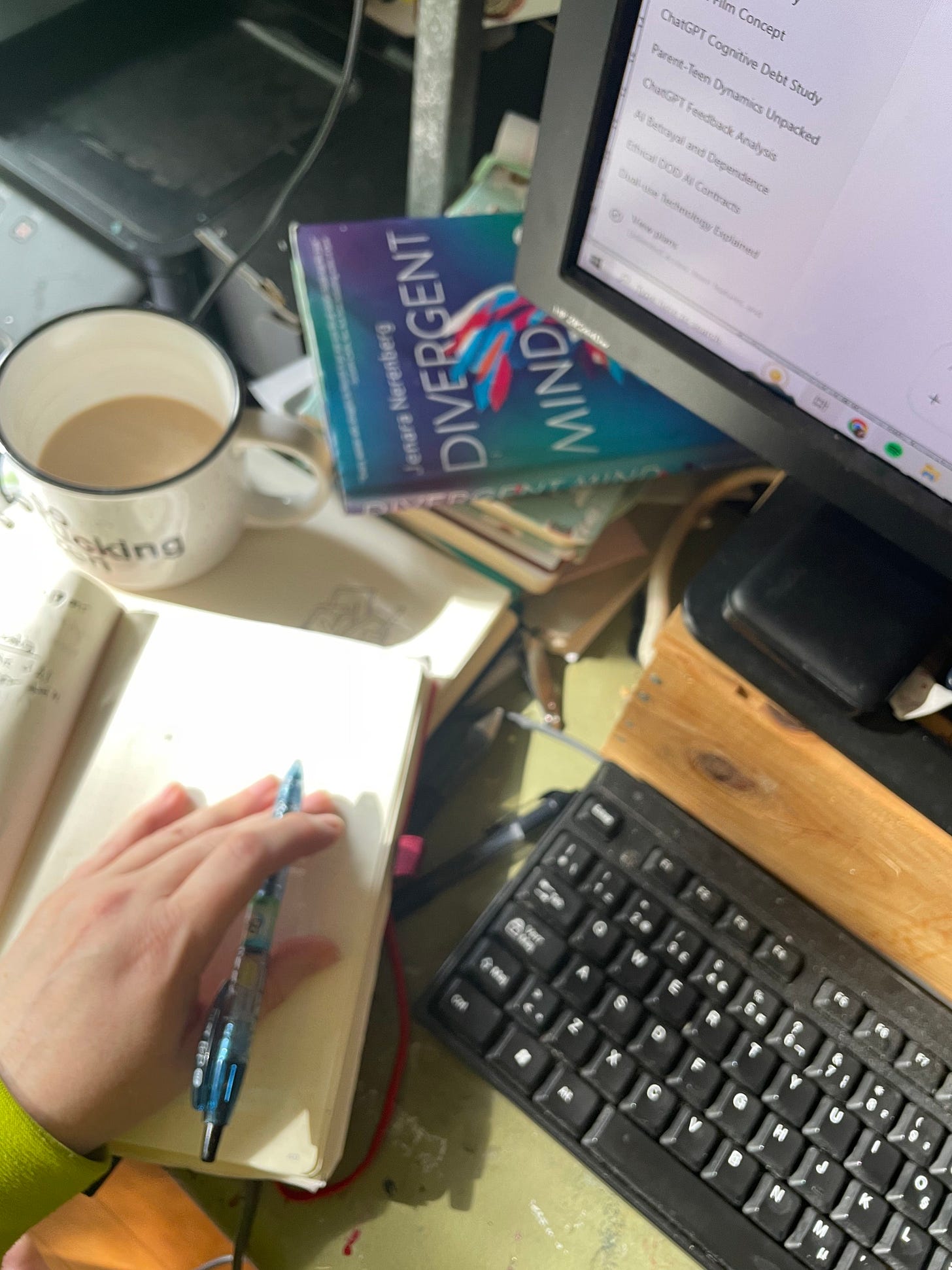 A cluttered workspace with a mug of coffee, an open notebook with a hand holding a pen, a computer keyboard, and the book “Divergent Mind” near a monitor showing an AI chat window.