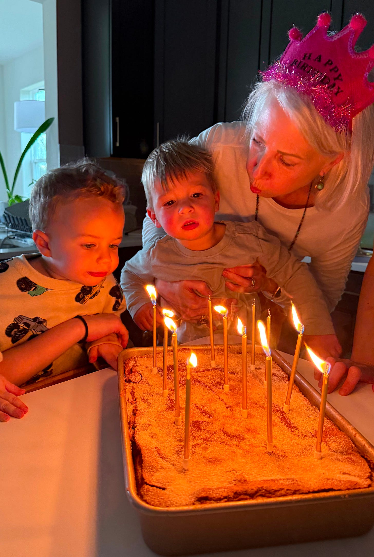 Ilona smiling and blowing out birthday candles with her two young grandsons at her 63rd birthday celebration.