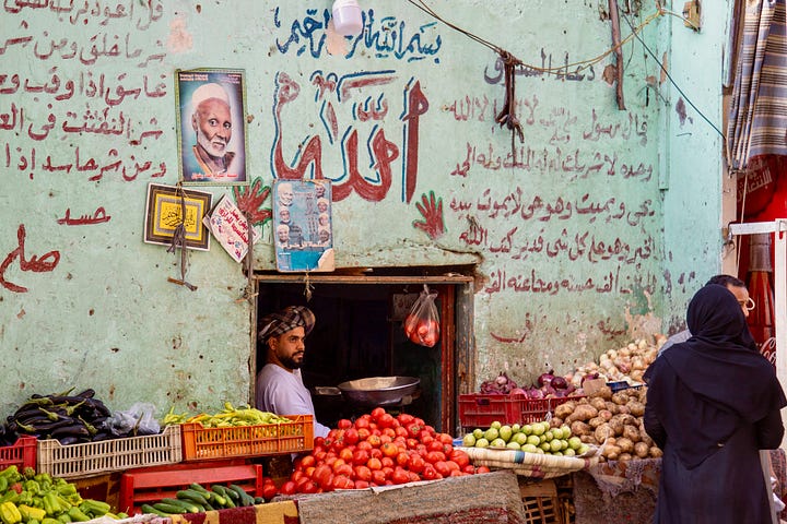 Top left: A narrow street in Luxor with apartment buildings, parked cars, and hanging textiles, with a Coptic church dome visible above the rooftops Top right: A woman in a black abaya buying fruit from a donkey-drawn cart loaded with oranges and bananas, parked in front of shuttered shopfronts Bottom right: A young man sitting inside a small vegetable stall framed by a green wall covered in Arabic calligraphy and religious posters, with piles of tomatoes, eggplant, and potatoes in the foreground Bottom left: A butcher in a blue plaid shirt bending over a cow's head on a cutting board, with a leg hanging from a hook and a Champions football poster on the red door behind him
