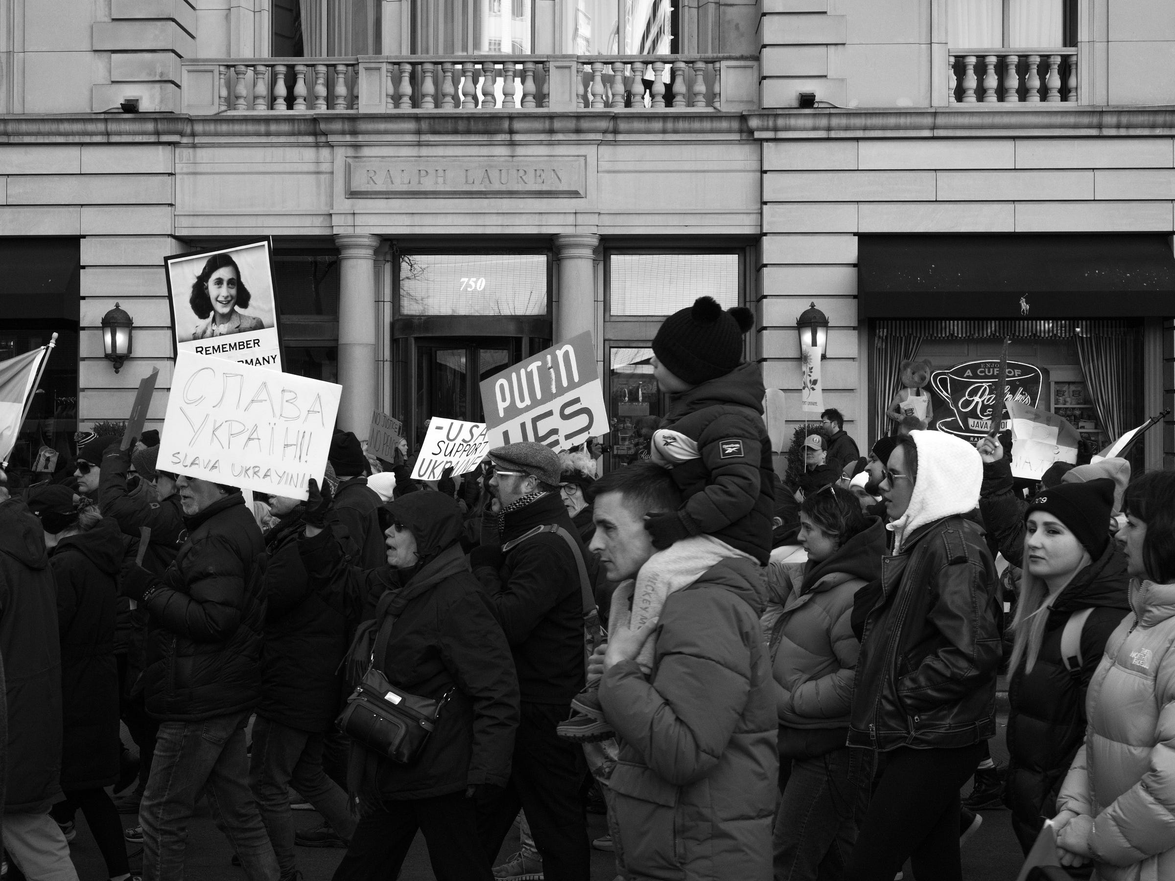 A black and white photograph captures a protest march in support of Ukraine. Protesters hold signs, including one that reads "Slava Ukraini!" and another featuring Anne Frank with the word "Remember." A child sits on a man's shoulders, bundled in winter clothing. The crowd moves past a Ralph Lauren storefront, with determined expressions and Ukrainian flags visible among them. A black and white photograph captures a protest march in support of Ukraine. Protesters hold signs, including one that reads "Slava Ukraini!" and another featuring Anne Frank with the word "Remember." A child sits on a man's shoulders, bundled in winter clothing. The crowd moves past a Ralph Lauren storefront, with determined expressions and Ukrainian flags visible among them.