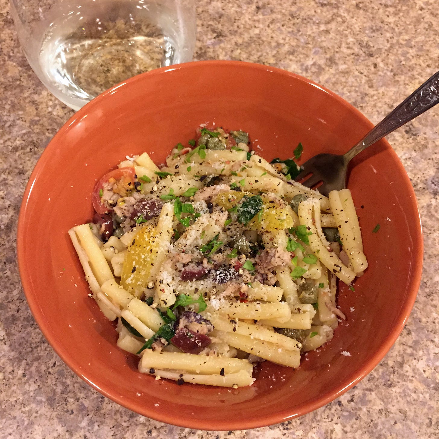 In an orange bowl is a serving of casarecce with flakes of tuna, pieces of tomato, arugula, and kalamata olives. Parsley, parmesan, and black pepper are sprinkled over the top. A small glass of white wine is just visible in the upper left corner.