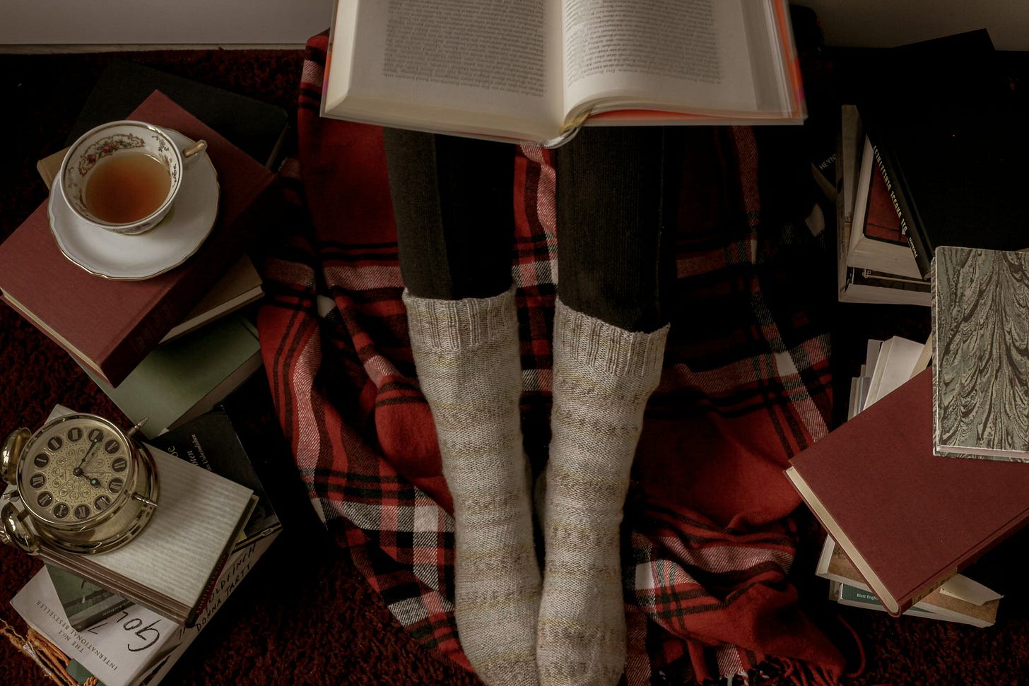 A person sitting reading, just calves in leggings with beige striped socks over their feet visible, resing on a red plaid blanket, an open book in their lap. To either side, books form stacks, one serving as a table for a cup of tea, and another holding a decorative alarm clock.