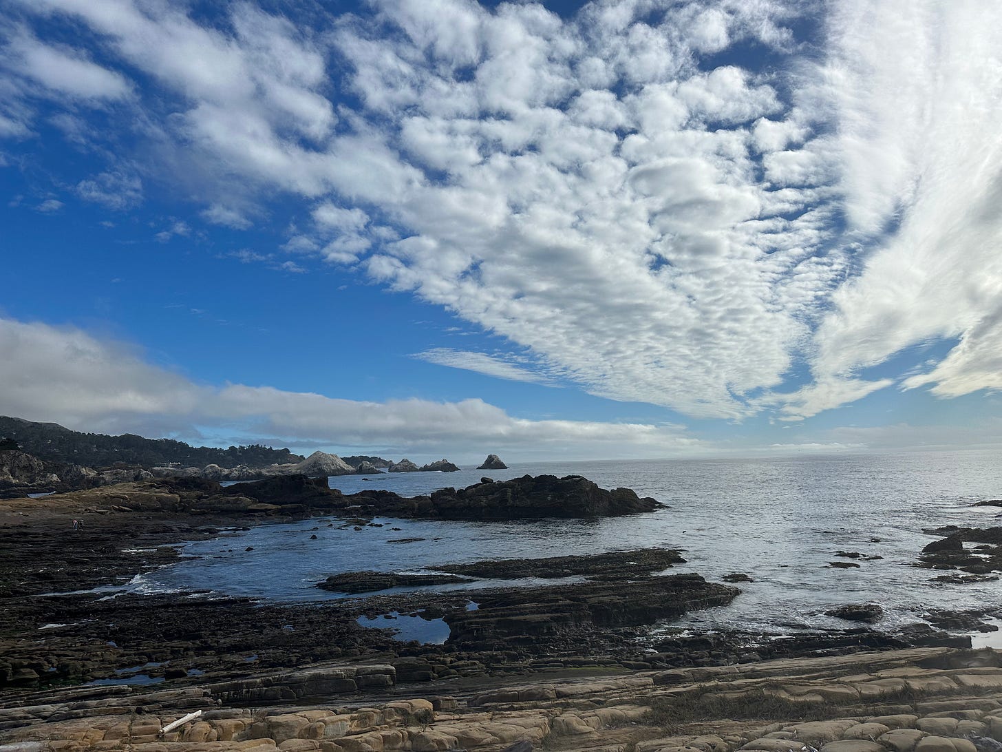 A blue sky streaked with clouds opens up over the ocean at low tide, spilling over rocky crags.