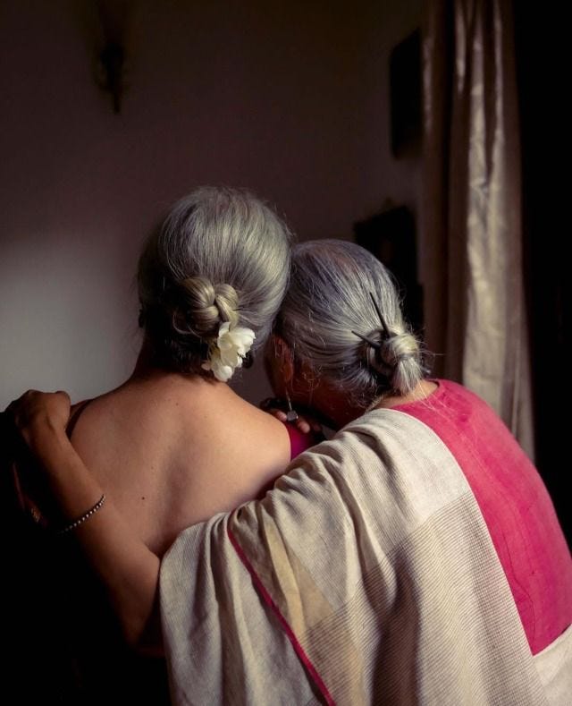 This may contain: two older women embracing each other in front of a window This may contain: two older women embracing each other in front of a window