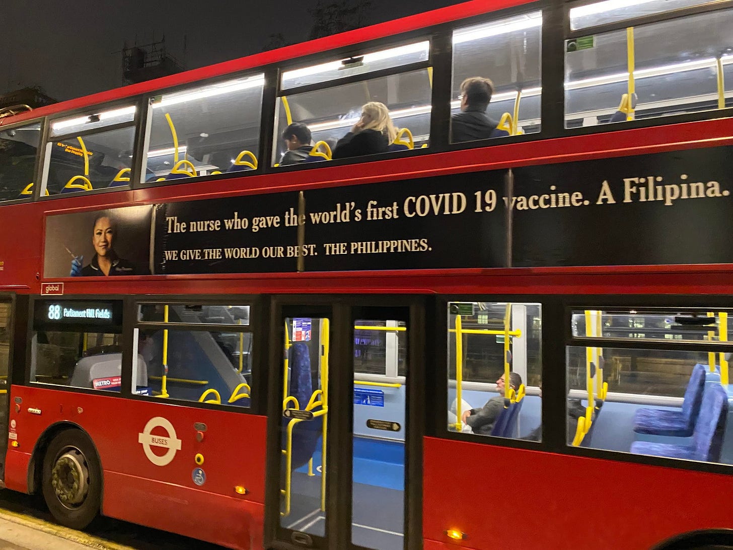 A red London-style double-decker bus with a side banner that has a picture of the Filipino-British nurse May Parsons and text that reads, "The nurse who gave the world's first COVID 19 vaccine. A Filipina." Below that is more text in all-caps that reads, "We give the world our best. The Philippines."