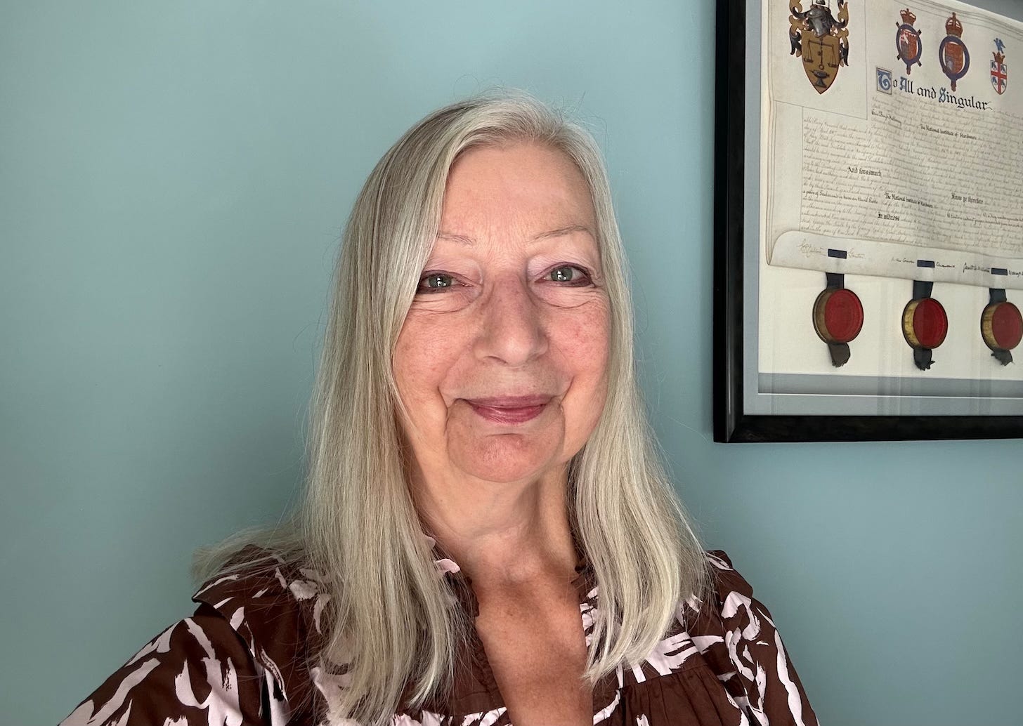 Head and shoulders image of a senior white woman with silver hair in front of a blue painted background smiling at the camera.