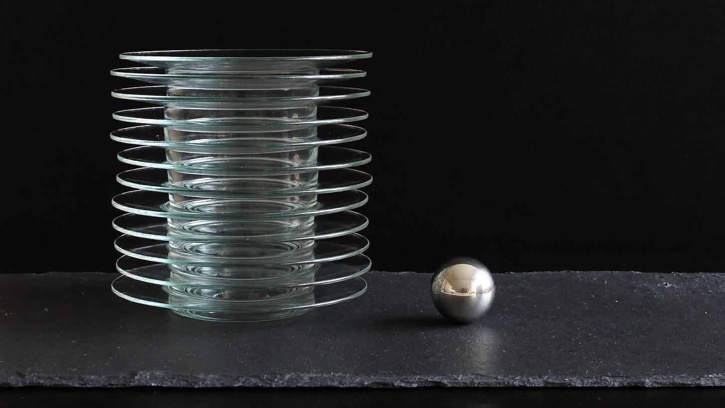 A tall stack of clear glass plates beside one small silver sphere on dark slate.