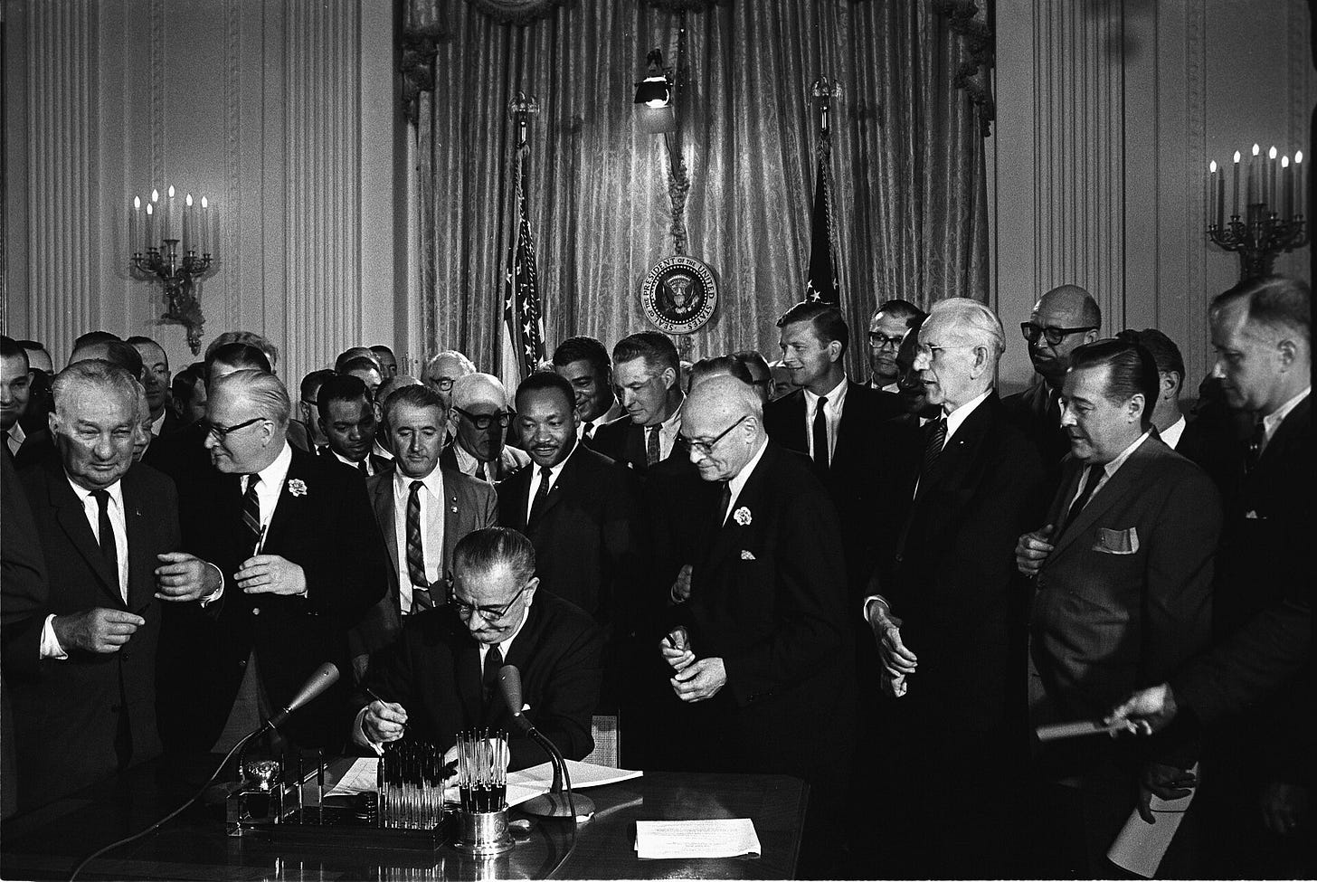 LBJ signs civil rights act in room full of white guys in suits; MLK Jr stands behind him.