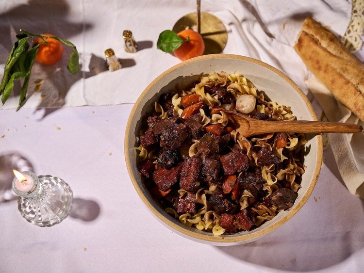 Bœuf Bourguignon served over buttered egg noodles in a large ceramic bowl, with chunks of slow-braised beef, carrots, and mushrooms, surrounded by baguette, clementines, and a lit candle on a white tablecloth.