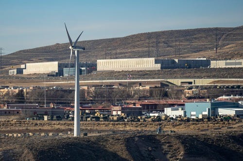 The NSA Utah Data Center on a hillside beyond a large wind turbine, with industrial buildings in the foreground.