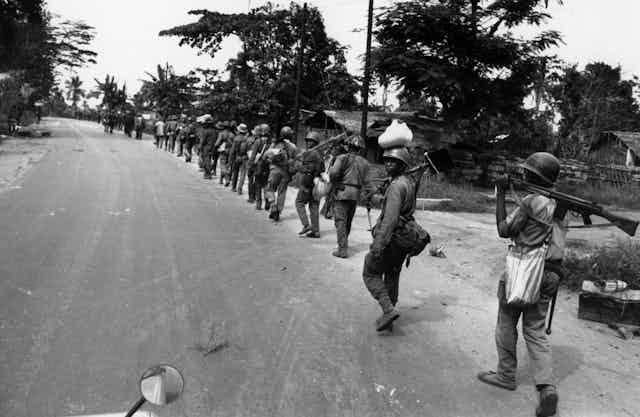 A black and white image of a long row of soldiers walking single file down a road, one looking back at the camera. A black and white image of a long row of soldiers walking single file down a road, one looking back at the camera.