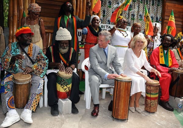 Charles sitting next to Camilla, wearing formal clothes, with drums in front of them. Several drummers and dancers in colorful clothing sit next to them. 