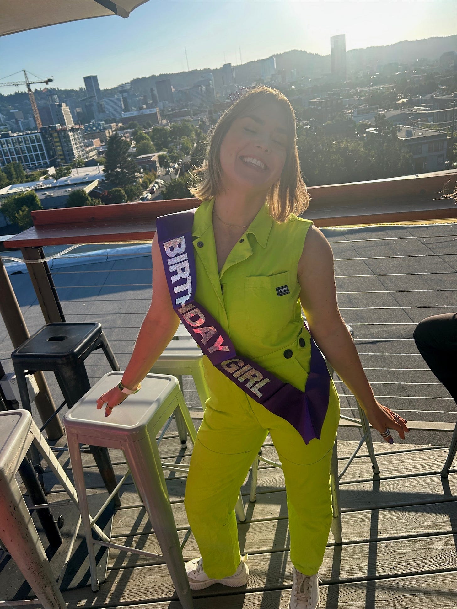 Cassidy wearing a neon yellow jumpsuit and a sash that reads "birthday girl," standing on a rooftop in Portland