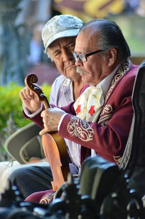 Um senhor mais velho afina um violino. Traja roupas enfeitadas, como um mariachi. Ao lado dele, um outro senhor, de boné e camisa meio aberta, o observa com atenção
