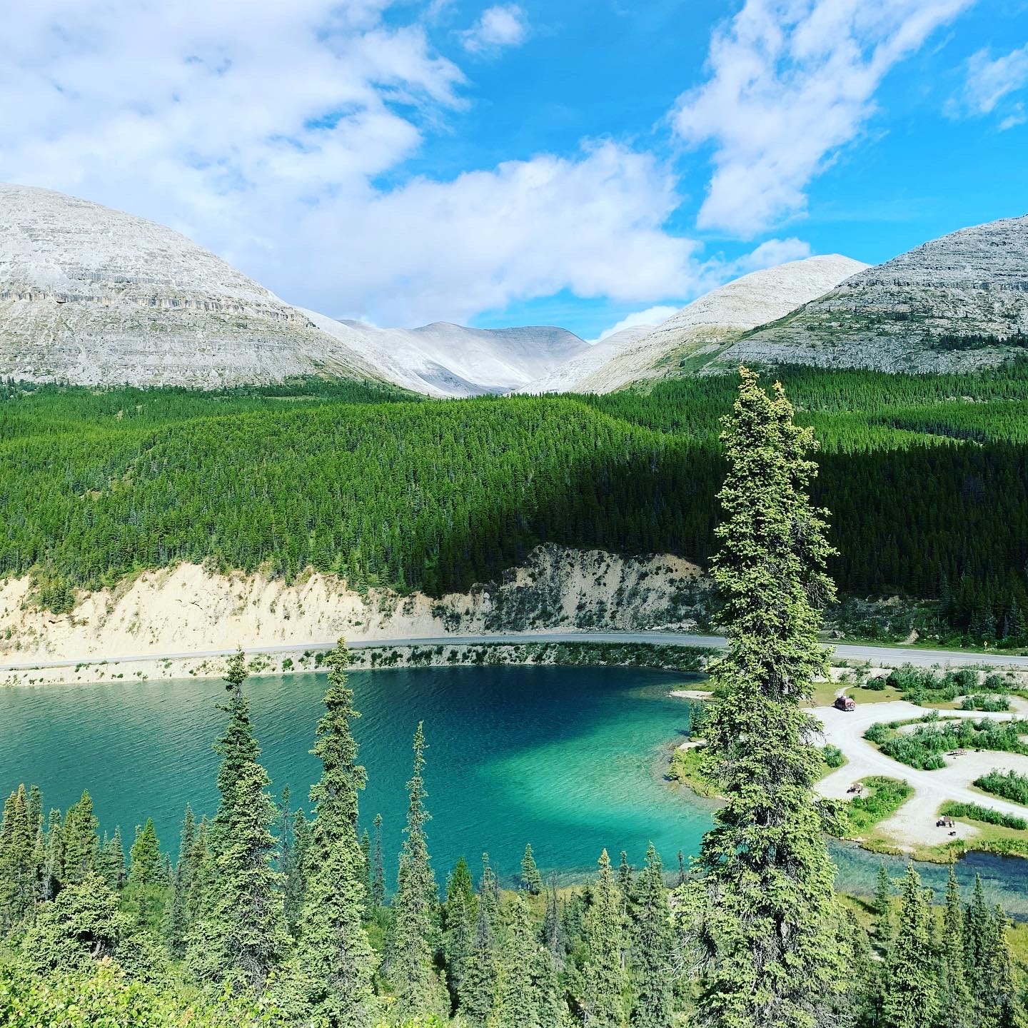In the foreground, a turquoise lake beneath a thicket of firs, in the background, another thicket of subalpine firs beneath a barer alpine layer and, above it, a blue sky In the foreground, a turquoise lake beneath a thicket of firs, in the background, another thicket of subalpine firs beneath a barer alpine layer and, above it, a blue sky